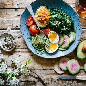 Ein rustikaler Holztisch mit einem blauen Salatteller mit Blattgemüse, Gurkenscheiben, Tomaten, Avocado, hart gekochtem Ei, geraspelten Karotten und Sonnenblumenkernen. Daneben liegen geschnittener Wassermelonenrettich, eine ganze Avocado, ein Ei und Blumen.