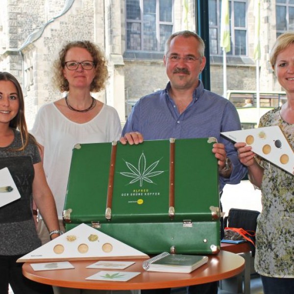 Vier Personen stehen in einem Raum und halten Aufklärungspakete und -materialien in der Hand, die aus einem grünen Koffer mit Cannabisblättern, runden Fächern und Broschüren bestehen, die auf einem Tisch vor ihnen liegen.