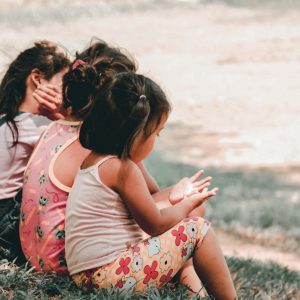 Drei kleine Kinder sitzen zusammen im Gras und schauen von der Kamera weg. Ein Kind bedeckt sein Gesicht, während die anderen auf ihre Hände schauen. Sie tragen legere Sommerkleidung und scheinen sich an einem sonnigen Tag im Freien aufzuhalten.