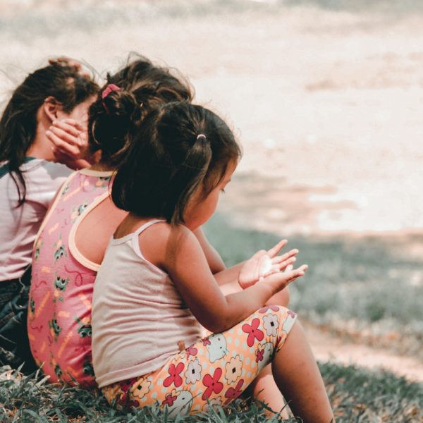 Drei kleine Kinder sitzen zusammen im Gras und schauen von der Kamera weg. Ein Kind bedeckt sein Gesicht, während die anderen auf ihre Hände schauen. Sie tragen legere Sommerkleidung und scheinen sich an einem sonnigen Tag im Freien aufzuhalten.
