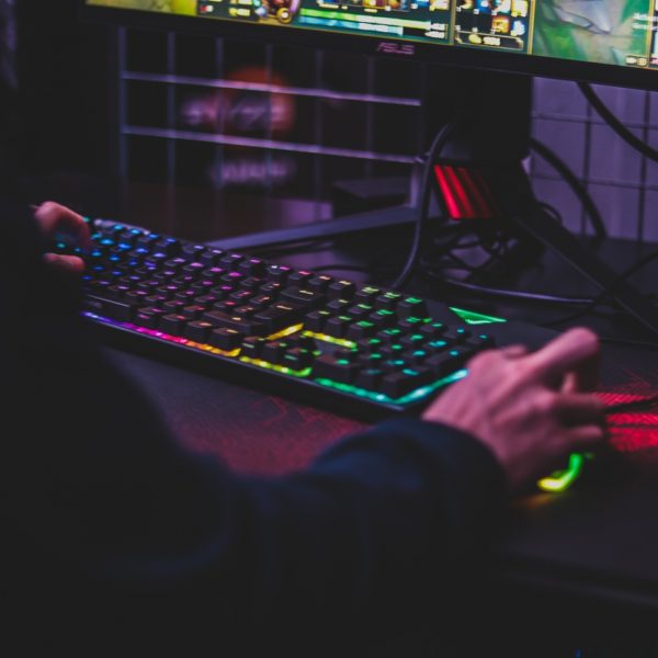 Eine Person benutzt eine bunte, beleuchtete Tastatur und Maus, während sie ein Videospiel auf einem Computermonitor spielt. Die Szene ist schwach beleuchtet und hebt die regenbogenfarbene Hintergrundbeleuchtung der Tastaturen hervor.