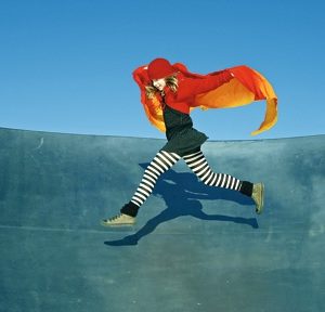 Eine Person, die einen rot-orangefarbenen Umhang, ein schwarzes Kleid und eine schwarz-weiß gestreifte Strumpfhose trägt, springt energisch vor einem blauen Himmel und einer gewölbten blauen Fläche und wirft einen Schatten auf den Boden.