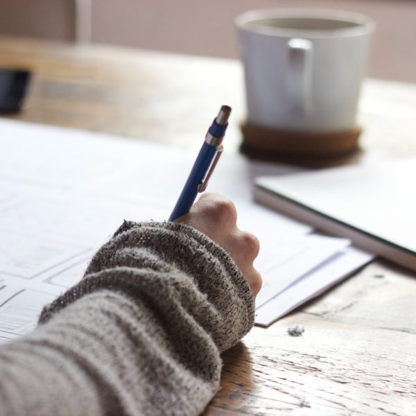 Eine Person in einem grauen Pullover schreibt an einem Holztisch auf Papieren, daneben stehen eine weiße Tasse und ein geschlossenes Notizbuch.