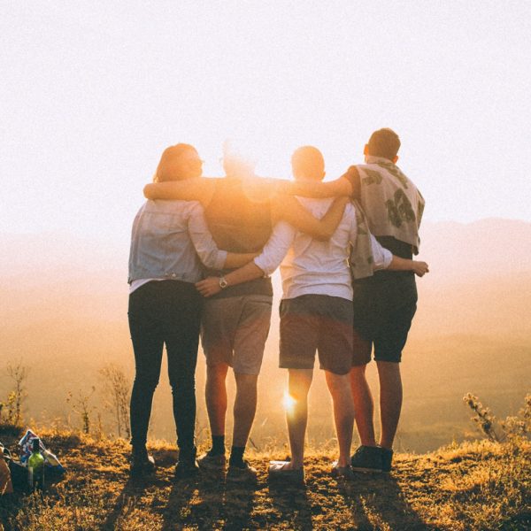 Vier Personen stehen Seite an Seite, die Arme umeinander gelegt, mit Blick auf einen malerischen Sonnenuntergang über Hügeln. Sie befinden sich im Freien, umgeben von Gras, und das Sonnenlicht scheint hell in den Hintergrund.