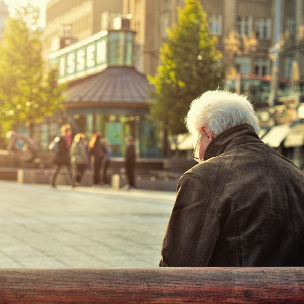 Ein älterer Mann mit weißem Haar sitzt allein auf einer Bank auf einem sonnenbeschienenen Platz in der Stadt, während im Hintergrund Menschen spazieren gehen und Bäume in der Nähe eines Gebäudes mit großen Fenstern zu sehen sind.