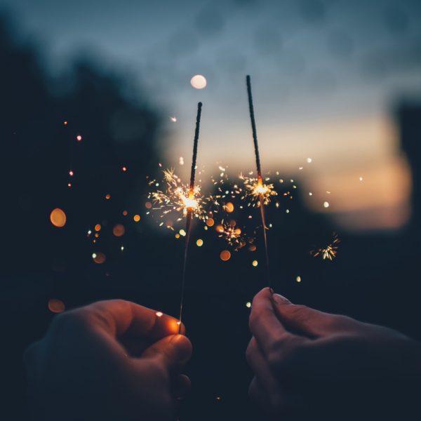 Zwei Hände, die Wunderkerzen eng aneinander halten, mit hellen Funken und Bokeh-Lichtern vor einem verschwommenen Hintergrund in der Dämmerung oder bei Nacht.