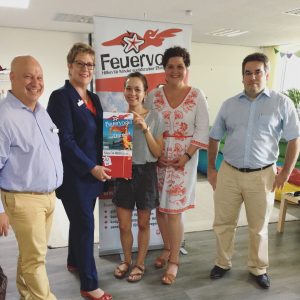 Fünf Personen stehen drinnen und lächeln in die Kamera; eine Frau hält eine Geschenktüte und eine Zeitschrift. Hinter ihnen ist ein Feuervogel-Banner mit deutschem Text und einem roten Vogellogo zu sehen. Die Umgebung sieht aus wie ein Büro oder ein Besprechungsraum.