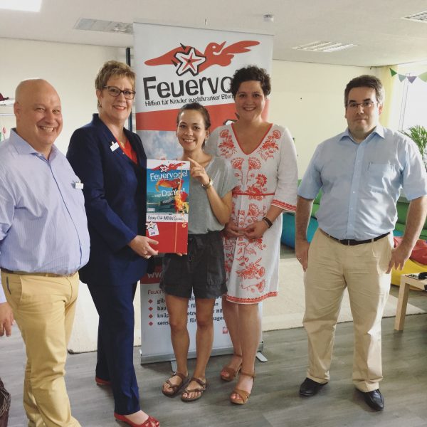 Fünf Personen stehen drinnen und lächeln in die Kamera; eine Frau hält eine Geschenktüte und eine Zeitschrift. Hinter ihnen ist ein Feuervogel-Banner mit deutschem Text und einem roten Vogellogo zu sehen. Die Umgebung sieht aus wie ein Büro oder ein Besprechungsraum.