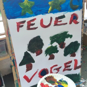 Ein Kind hält einen Pappteller mit roten, grünen, gelben und blauen Farbklecksen in der Hand und malt ein Plakat mit dem deutschen Wort FEUER VOGEL und Bildern von Sternen, einer Mondsichel und grünen Bäumen.
