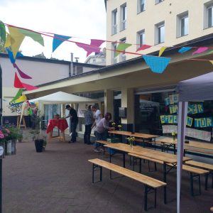 Veranstaltung im Freien mit bunten Wimpeln, Holzbänken, Tischen mit Blumen und Menschen, die vor einem Gebäude Dekorationen arrangieren. Schilder und ein überdachtes Zelt sind zu sehen und schaffen eine festliche Atmosphäre.