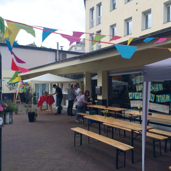 Veranstaltung im Freien mit bunten Wimpeln, Holzbänken, Tischen mit Blumen und Menschen, die vor einem Gebäude Dekorationen arrangieren. Schilder und ein überdachtes Zelt sind zu sehen und schaffen eine festliche Atmosphäre.