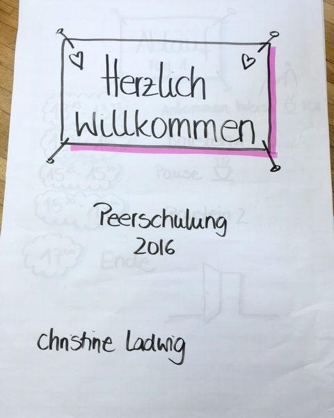 Auf einem handgeschriebenen Schild auf weißem Papier steht: Herzlich willkommen, Peerschulung 2016, und Christine Ludwig, mit kleinen Herzchen und einem rosa Rand um den oberen Text.