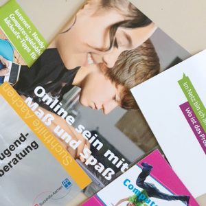 Auf einem Tisch liegen ein paar bunte Broschüren übereinander. Eine zeigt zwei Teenager, die einen Laptop benutzen, und den deutschen Text Online sein mit Maß und Spaß. Andere Broschüren enthalten Texte über Jugendberatung und Computerspiele.