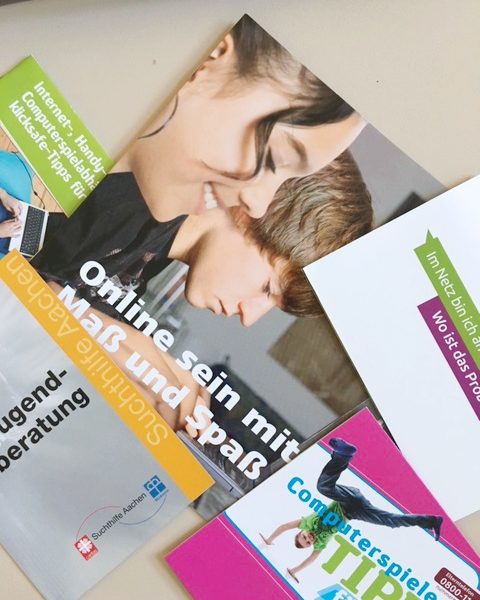 Auf einem Tisch liegen ein paar bunte Broschüren übereinander. Eine zeigt zwei Teenager, die einen Laptop benutzen, und den deutschen Text Online sein mit Maß und Spaß. Andere Broschüren enthalten Texte über Jugendberatung und Computerspiele.