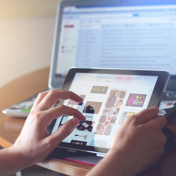 Eine Person benutzt ein Tablet und blättert in Bildern, während im Hintergrund auf einem runden Holztisch ein Laptop mit unscharfem Bildschirm steht. Die Umgebung scheint zwanglos und gut beleuchtet zu sein.
