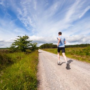Eine Person in Sportkleidung joggt allein auf einem Schotterweg durch eine ländliche Landschaft unter einem strahlend blauen Himmel mit Wolkenfetzen, umgeben von Gras und Bäumen.