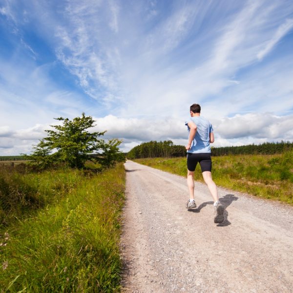Eine Person in Sportkleidung joggt allein auf einem Schotterweg durch eine ländliche Landschaft unter einem strahlend blauen Himmel mit Wolkenfetzen, umgeben von Gras und Bäumen.