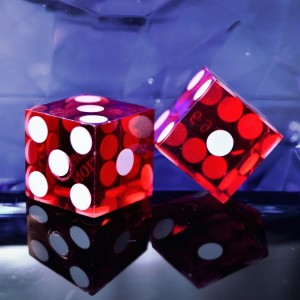Zwei durchsichtige rote Würfel mit weißen Punkten liegen auf einer reflektierenden Oberfläche vor einem blauen, strukturierten Hintergrund. Ein Würfel liegt flach, während der andere auf die Kante gekippt ist.
