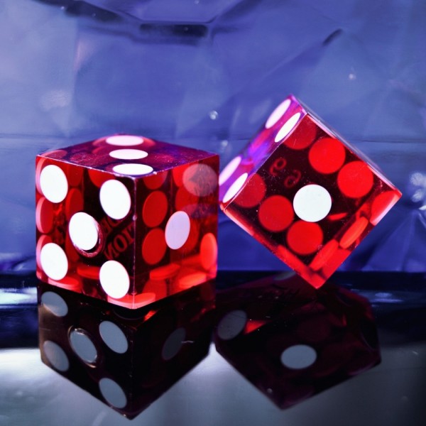 Zwei durchsichtige rote Würfel mit weißen Punkten liegen auf einer reflektierenden Oberfläche vor einem blauen, strukturierten Hintergrund. Ein Würfel liegt flach, während der andere auf die Kante gekippt ist.