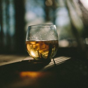 Eine Nahaufnahme eines Glases, das mit einem goldenen Getränk gefüllt ist, steht auf einer hölzernen Oberfläche in sanftem, gesprenkeltem Sonnenlicht, mit einem verschwommenen Waldhintergrund, der eine ruhige, beschauliche Atmosphäre schafft.