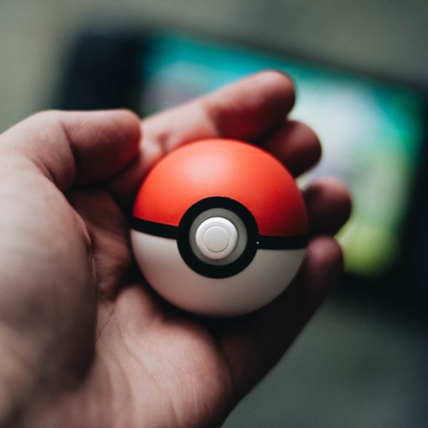 Eine Hand, die einen Pokéball hält, mit einem unscharfen Hintergrund, der ein mobiles Gerät oder eine Spielkonsole zu sein scheint.
