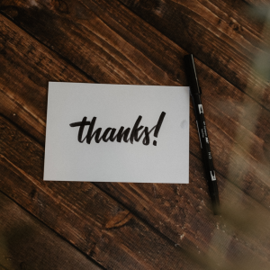 Eine weiße Karte, auf der in fetter schwarzer Schrift "Danke!" geschrieben steht, liegt auf einer hölzernen Unterlage neben einem schwarzen Stift.