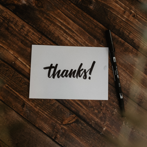 Eine weiße Karte, auf der in fetter schwarzer Schrift "Danke!" geschrieben steht, liegt auf einer hölzernen Unterlage neben einem schwarzen Stift.