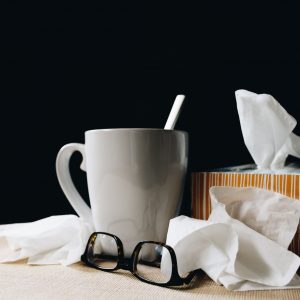 Ein weißer Becher mit Löffel, eine gestreifte Taschentuchbox, lose Taschentücher und eine Brille stehen auf einem Tisch vor einem dunklen Hintergrund.