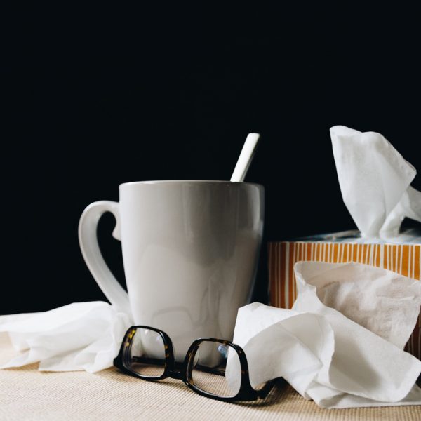 Ein weißer Becher mit Löffel, eine gestreifte Taschentuchbox, lose Taschentücher und eine Brille stehen auf einem Tisch vor einem dunklen Hintergrund.