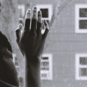 Eine Hand mit einem Ring berührt sanft ein regennasses Fenster, während draußen ein unscharfes Gebäude und ein kahler Baum zu sehen sind. Die Szene wirkt ruhig und kontemplativ.