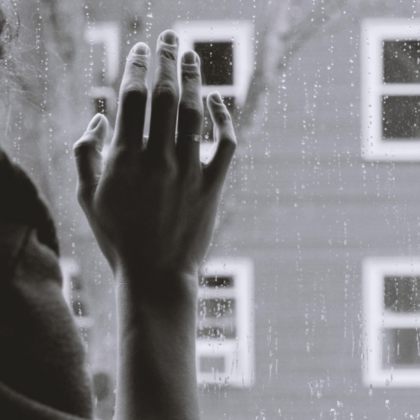 Eine Hand mit einem Ring berührt sanft ein regennasses Fenster, während draußen ein unscharfes Gebäude und ein kahler Baum zu sehen sind. Die Szene wirkt ruhig und kontemplativ.