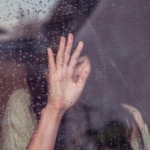 Eine Person drückt ihre Hand gegen ein regennasses Fenster, wobei Wassertropfen auf das Glas fallen und ihr Gesicht teilweise verdeckt ist, was eine düstere und nachdenkliche Stimmung erzeugt.