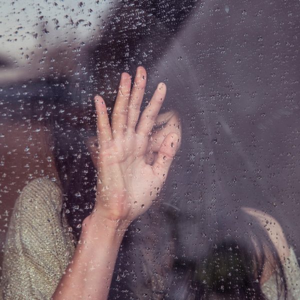 Eine Person drückt ihre Hand gegen ein regennasses Fenster, wobei Wassertropfen auf das Glas fallen und ihr Gesicht teilweise verdeckt ist, was eine düstere und nachdenkliche Stimmung erzeugt.