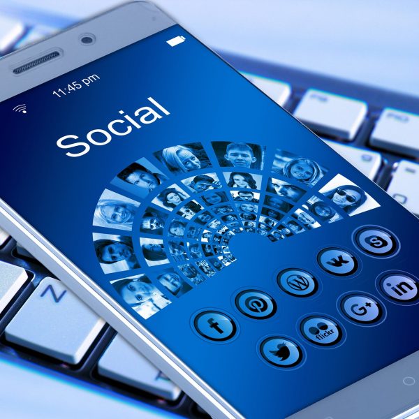 Ein Smartphone, das einen sozialen Bildschirm mit verschiedenen Social-Media-Symbolen und Profilfotos anzeigt und auf einer Computertastatur liegt. Das Telefon zeigt Symbole für Facebook, Twitter, Pinterest, WhatsApp, LinkedIn, Google+ und andere an.