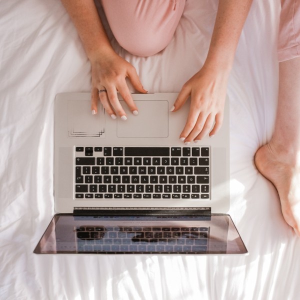 Eine Person im rosa Schlafanzug sitzt im Schneidersitz auf einem weißen Bett und tippt auf einem silbernen Laptop. Nur ihre Hände, Beine und ein Teil des Laptops sind sichtbar.