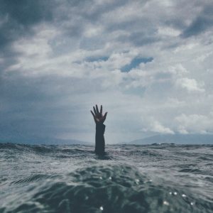 Eine einzelne Hand ragt unter einem wolkenverhangenen Himmel aus dem rauen Wasser des Ozeans und deutet auf eine Person hin, die sich inmitten der stürmischen Wellen abmüht oder um Hilfe ruft.