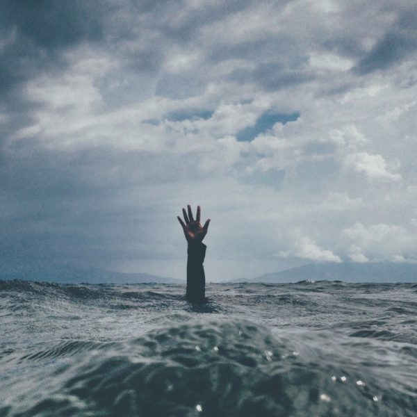 Eine einzelne Hand ragt unter einem wolkenverhangenen Himmel aus dem rauen Wasser des Ozeans und deutet auf eine Person hin, die sich inmitten der stürmischen Wellen abmüht oder um Hilfe ruft.