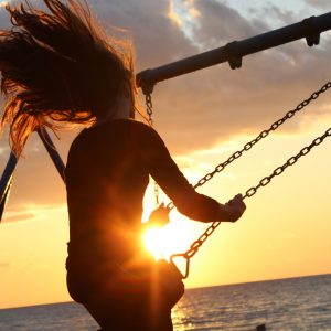 Eine Person mit langen Haaren schaukelt bei Sonnenuntergang auf einer Spielplatzschaukel am Meer, wobei die Sonne durch die Schaukel scheint und ihr Haar im Wind weht.
