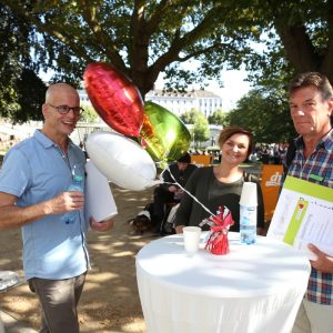 Drei Personen stehen bei einer Veranstaltung im Park um einen hohen Tisch herum, in der Hand Luftballons und Getränke. Bäume spenden Schatten und im Hintergrund sind weitere Menschen und Tische zu sehen.