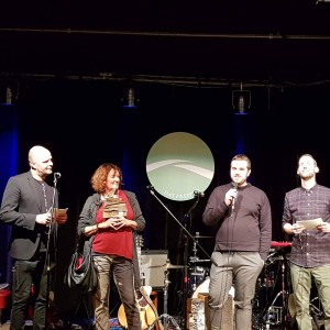 Vier Personen stehen auf einer Bühne mit Mikrofonen, sprechen und halten Papiere in der Hand. Hinter ihnen sind Musikinstrumente und Geräte zu sehen, und im Hintergrund hängt ein rundes Schild mit Text im Bühnenlicht.
