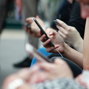 Eine Gruppe eng beieinander sitzender Personen, die jeweils ein Smartphone halten und benutzen. Der Fokus liegt auf ihren Händen und Telefonen, während der Hintergrund und die Gesichter unscharf sind.