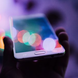 Eine Hand hält ein weißes Smartphone vor einem bunten, verschwommenen Hintergrund aus Bokeh-Lichtern, der eine verträumte und lebendige Atmosphäre schafft. Der Bildschirm des Telefons reflektiert einige der hellen Lichter.