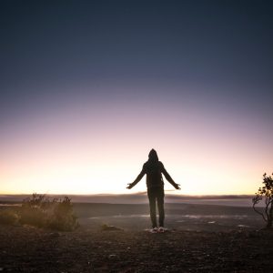 Eine Person im Kapuzenpulli steht mit leicht ausgestreckten Armen auf einer Bergkuppe bei Sonnenaufgang oder Sonnenuntergang, vor einem bunten Himmel mit schwachem Licht am Horizont und minimaler Vegetation in der Nähe.