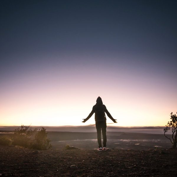 Eine Person im Kapuzenpulli steht mit leicht ausgestreckten Armen auf einer Bergkuppe bei Sonnenaufgang oder Sonnenuntergang, vor einem bunten Himmel mit schwachem Licht am Horizont und minimaler Vegetation in der Nähe.