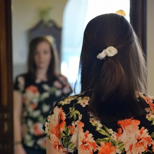 Eine Frau mit braunem Haar, das mit einem weißen Haargummi zurückgebunden ist, steht vor einem Spiegel. Sie trägt ein schwarzes Hemd mit einem bunten Blumenmuster und ihr Spiegelbild ist zu sehen.