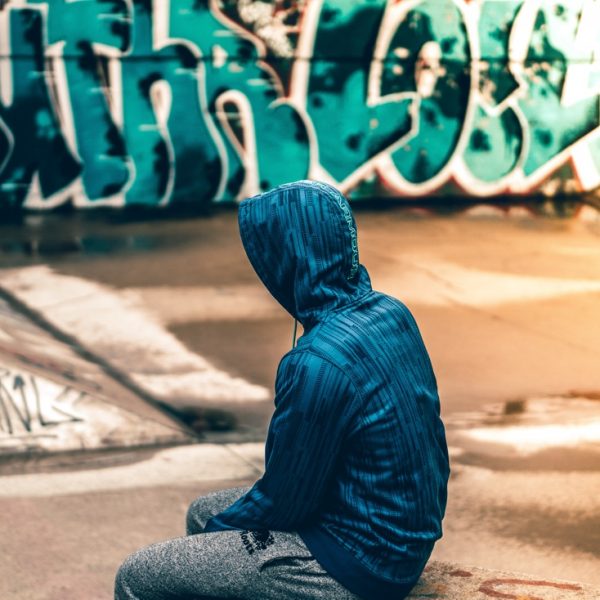 Eine Person, die einen blau gestreiften Kapuzenpulli und eine graue Hose trägt, sitzt auf einer Betonfläche vor einer mit bunten Graffiti beschmierten Wand. Die Person blickt von der Kamera weg.