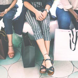 Drei Frauen sitzen nebeneinander mit Einkaufstüten zu ihren Füßen. Nur ihre Beine und Hände sind zu sehen. Sie sind leger gekleidet und scheinen sich nach dem Einkaufen zu entspannen.