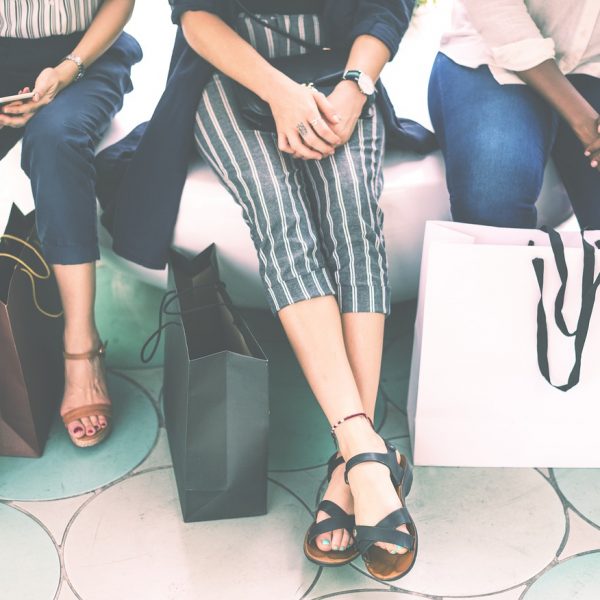Drei Frauen sitzen nebeneinander mit Einkaufstüten zu ihren Füßen. Nur ihre Beine und Hände sind zu sehen. Sie sind leger gekleidet und scheinen sich nach dem Einkaufen zu entspannen.