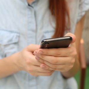 Eine Person mit langen Haaren, die ein hellblaues kurzärmeliges Hemd trägt, hält und benutzt mit beiden Händen ein Smartphone. Der Hintergrund ist unscharf und zeigt etwas Grün und einen Holzstuhl.