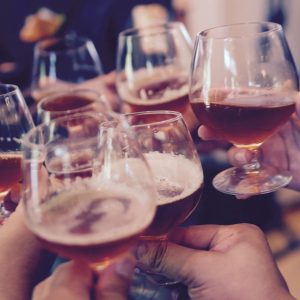 Eine Gruppe von Menschen stößt mit Gläsern an, die mit bernsteinfarbenen Getränken gefüllt sind, und feiert. Die Szene ist lebhaft, und mehrere Hände halten runde Gläser, was auf ein geselliges Beisammensein oder eine Feier hindeutet.